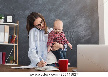 Happy Mother Working At Office With Her Baby. Young Woman Talking On Phone And Writing Notes While S