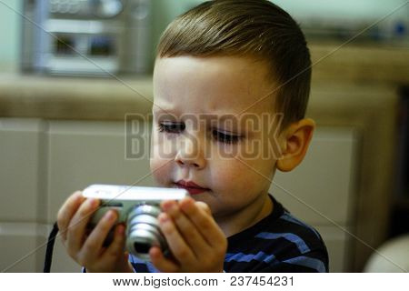 A Small Boy Holds A Camera In His Hands And Stares At The Screen With Comprehension. Memories, Pictu
