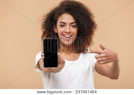 Portrait of a smiling young african woman poinitng finger at blank screen mobile phone isolated over beige background