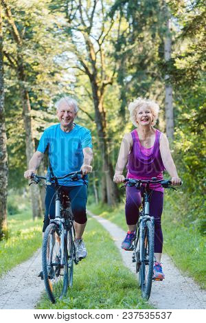 Full length of a happy and active senior couple wearing cool fitness outfits while riding bicycles outdoors in the park