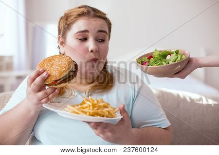 Think Twice. Obese Young Woman Choosing Between Hamburger And Salad While Sitting On The Couch