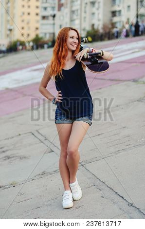 Redhead Slim Girl In Jeans Shorts And Black T Shirt Posing With Skateboard In Urban Sciene.