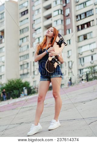 Redhead Slim Girl In Jeans Shorts And Black T Shirt Posing With Skateboard In Urban Sciene.