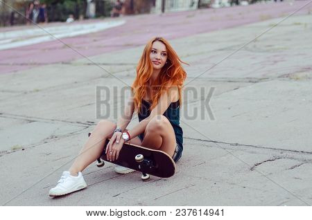 Redhead Slim Girl In Jeans Shorts And Black T Shirt Posing With Skateboard In Urban Sciene.