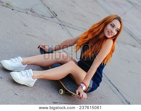 Redhead Slim Girl In Jeans Shorts And Black T Shirt Posing With Skateboard In Urban Sciene.