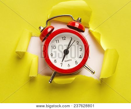 Red Alarm Clock Showing Up From Torn Yellow Paper At White Background. Time Is Our Most Precious Res