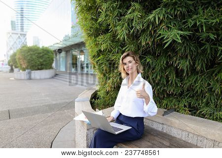 Caucasian Female Manager Working With Laptop And Papers In Open Air Near Green Plant, Showing Thumbs