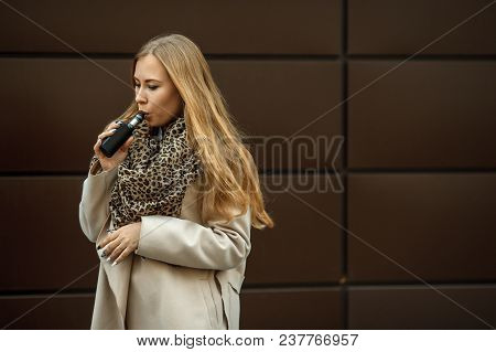 Vape Teenager. A Young Cute White Girl In Warm Scarf Is Vaping An Electronic Cigarette On The Street