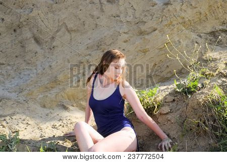 Beautiful Blonde In A Blue Bathing Suit Sits On A Steep Bank. Coast Of Clay And Sand.