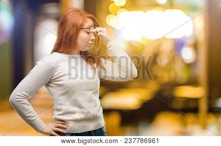 Beautiful young redhead woman with sleepy expression, being overworked and tired, rubbes nose because of weariness at night