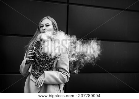 Vape Teenager. A Young Cute White Girl In Warm Scarf Is Vaping An Electronic Cigarette On The Street