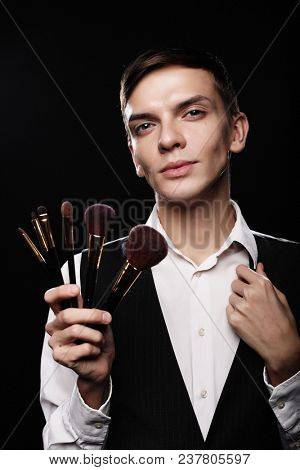 Ready to do make up. Young male make up artist posing with make-up brushes on a dark background