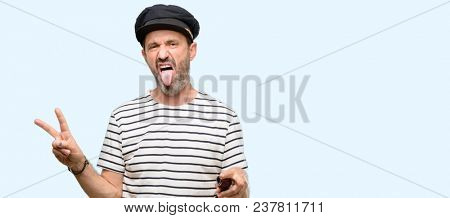 Sailor captain man smoking a tobacco pipe looking at camera showing tong and making victory sign with fingers isolated over blue background