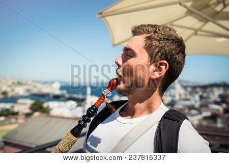 A Young Handsome Man With Beard Is Smoking A Hookah On The Terrace With View On The Cityscape