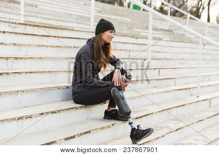 Photo of atlethic disabled woman in sportswear with prosthetic leg, sitting at the stairs outdoor and holding thermos cup