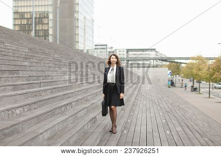 Caucasian Businesswoman Standing On Stairs With Bag And High Buildings In Background. Concept Of Pro