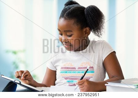 Close Up Portrait Of Cute Little African Girl Doing Home Work At Desk. Ponytailed Kid Typing On Digi