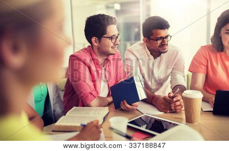 education, high school, people and technology concept - group of international students sitting at table with tablet pc computers, books and notebooks at university