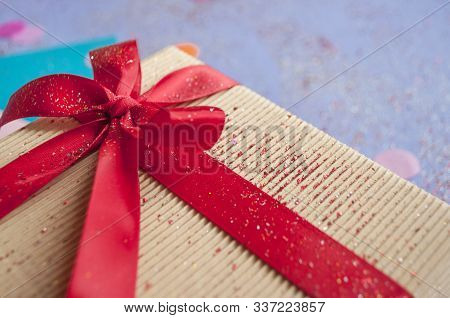 Close Up Of Giftbox Tied With Bright Red Color Ribbon On Purple Background With Sparklets And Confet