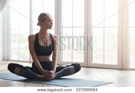 Pensive Smiling Girl Stretching Legs Before Work Out At Studio, Sitting On Yoga Mat, Looking At Wind