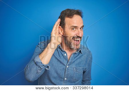 Handsome middle age senior man with grey hair over isolated blue background smiling with hand over ear listening an hearing to rumor or gossip. Deafness concept.