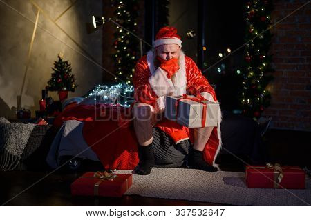 Bad Evil Santa Claus With A Red Beard Sits On The Bed And Collects Gifts On Christmas Eve. A Man In 