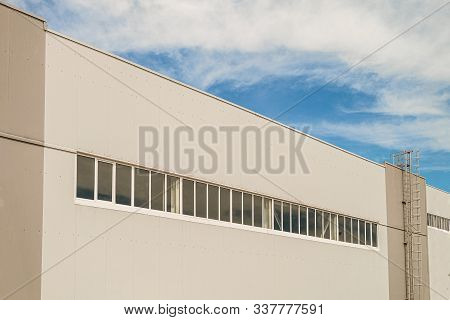 Facade Of A Long Multi-span Industrial Building Under A Blue Cloudy Sky. Gray Wall And Tape Glazing 