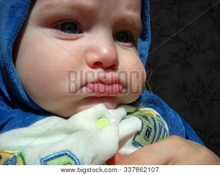 Baby 6 Months Old On Mothers Hands. The Boy Is Dressed In A Jumpsuit And A Blue Hood. A Child With T
