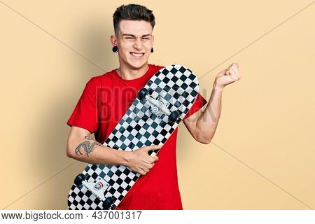 Young caucasian boy with ears dilation holding skate screaming proud, celebrating victory and success very excited with raised arm 
