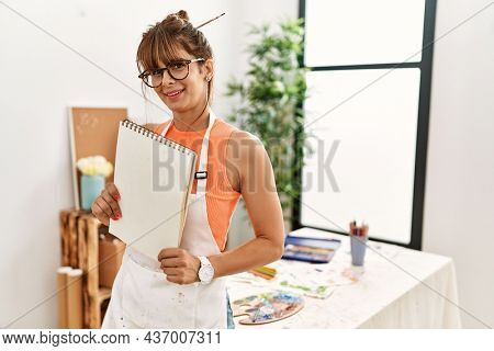 Young hispanic woman smiling confident holding notebook at art studio