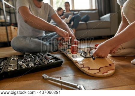 Crop Close Up Of Father And Son Sit On Floor Use Tools Repair Skateboard At Home Together. Caring Da