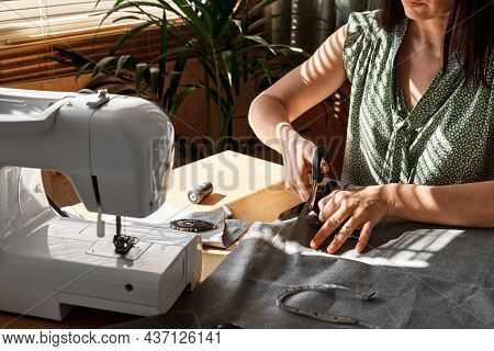 Woman Cutting Out A Pattern Paper In Linen Fabric. Seamstress Sewing On The Sewing Machine In Small 