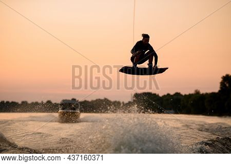 Active Man Flying Over Splashing Wave On Wakeboard Holding On To The Rope. Water Sports Activity.