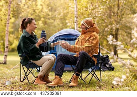 camping, tourism and travel concept - happy couple with thermos drinking tea to cup at tent camp