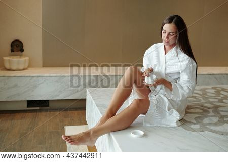 Young beautiful woman in white bathrobe applying moisturizing cream on her legs after hamam bath