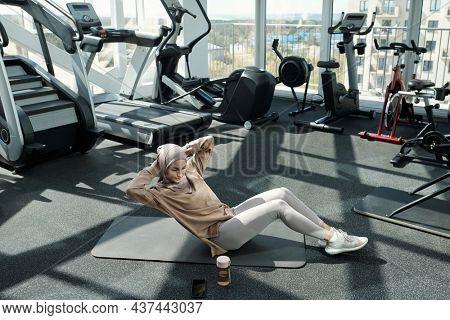 Young pretty sportswoman doing sit-ups with side turns while sitting on mat and keeping her hands behind head