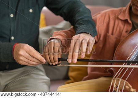 Hands of music teacher and his pupil playing cello while young man holding fiddlestick and helping little boy to glide it