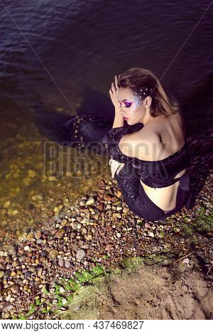 Mermaid Near Sea Shore On Rocks While Wearing Artistic Makeup With Facial Strassses And Black Shiny 
