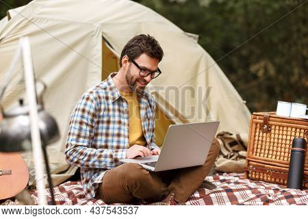 Smiling Man Sitting Near Tent With Laptop Computer And Working Remotely While Camping In Nature
