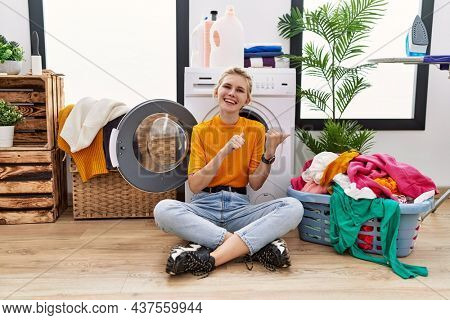 Young blonde woman doing laundry sitting by washing machine pointing to the back behind with hand and thumbs up, smiling confident 