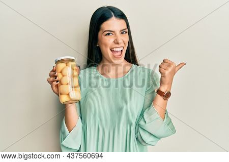 Young hispanic woman holding bottle with boiled potatoes pointing thumb up to the side smiling happy with open mouth 
