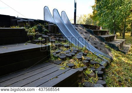 Double Shiny Stainless Steel Slide On The Playground. A Hill Lined With Wooden Logs Used As Palisade