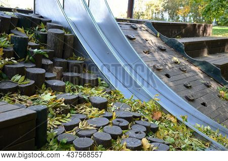 Double Shiny Stainless Steel Slide On The Playground. A Hill Lined With Wooden Logs Used As Palisade