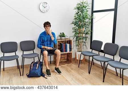 Young hispanic man sitting at waiting room with folder covering mouth with hand, shocked and afraid for mistake. surprised expression 