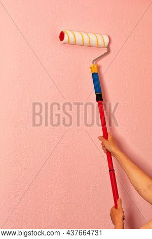Female Hands Of The Painter Hold A Paint Roller On A Long Telescopic Rod Against The Background Of A
