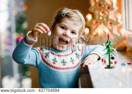 Litte Toddler Girl Sitting By Window And Decorating Small Glass Christmas Tree With Tiny Xmas Toys. 