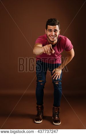 Young, Joyful Hipster Guy Points His Index Fingers In Front Of Him While Isolated Brown Background