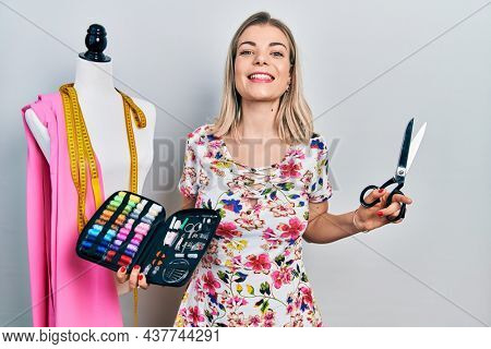 Beautiful caucasian woman dressmaker designer holding scissors and sewing kit smiling and laughing hard out loud because funny crazy joke. 