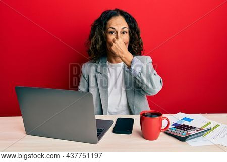 Beautiful middle age woman working at the office drinking a cup of coffee smelling something stinky and disgusting, intolerable smell, holding breath with fingers on nose. bad smell 