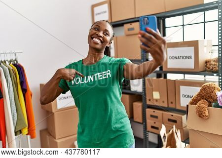 Young african american woman pointing with finger to volunteer tshirt make selfie by the smartphone at charity center.